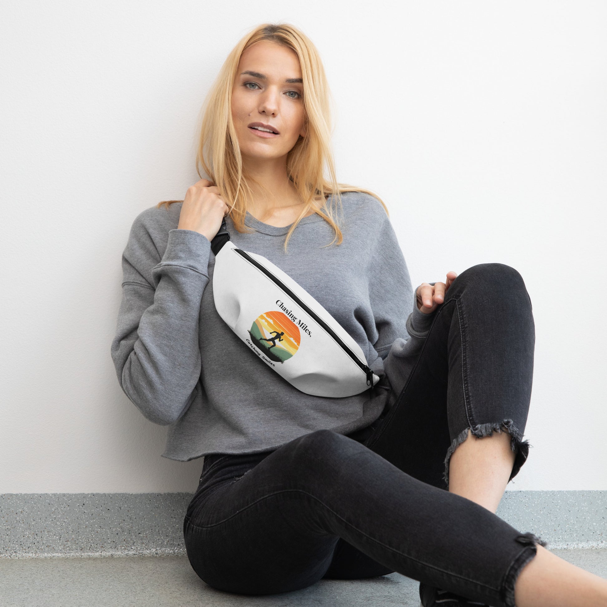 Woman sitting on the floor wearing a gray sweatshirt and black pants, holding a white fanny pack with a design.