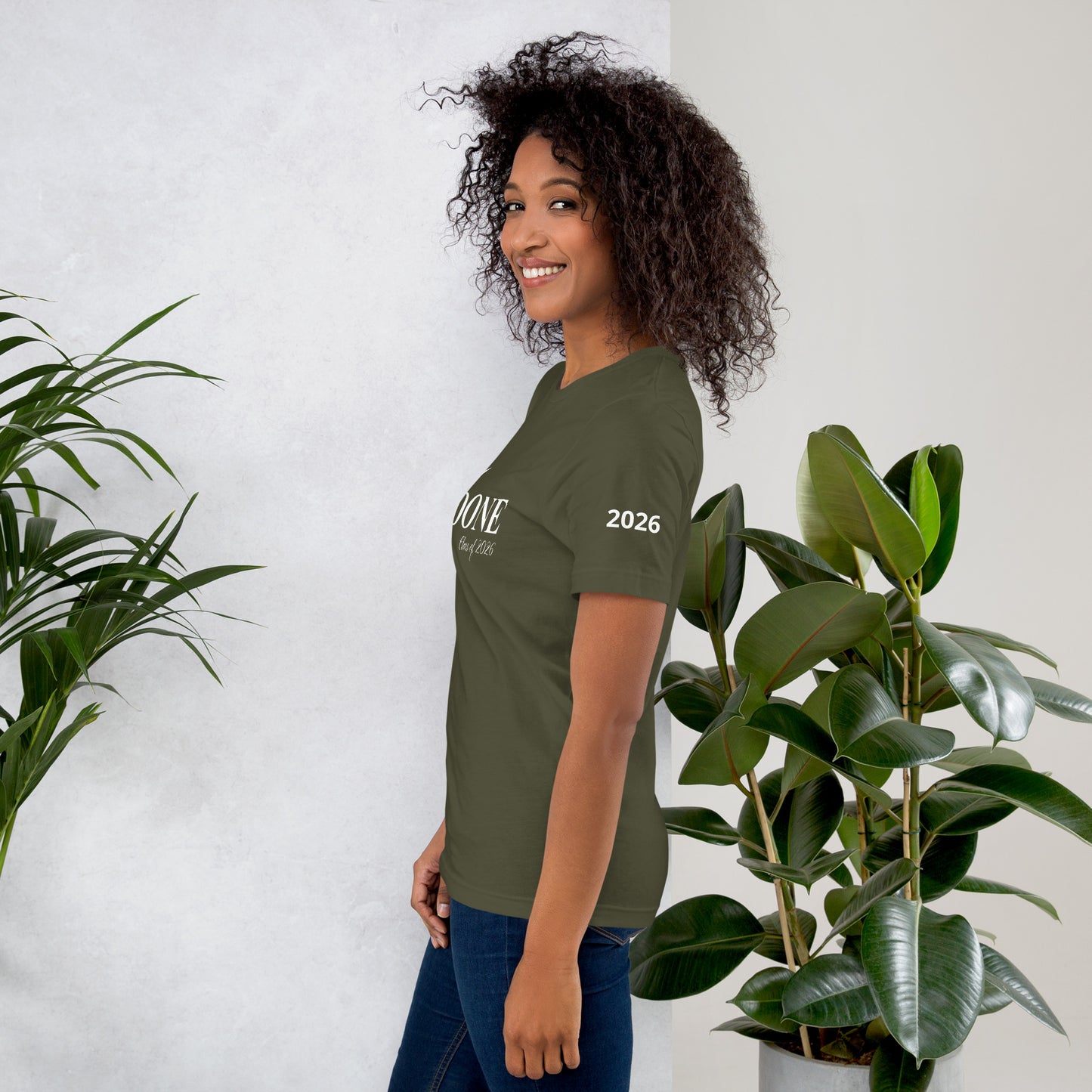 Woman wearing a green t-shirt with text, standing between two potted plants against a white wall.