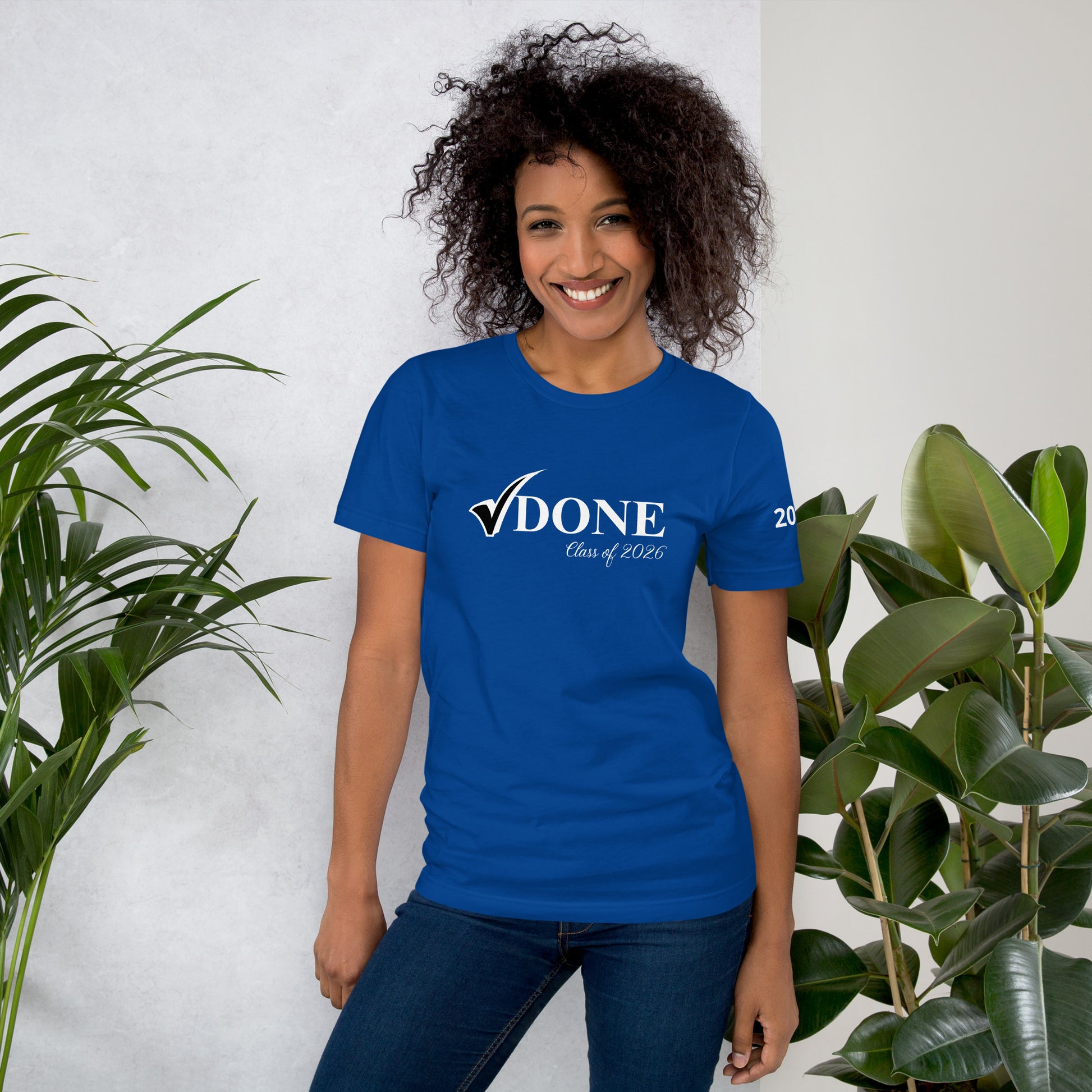 Woman wearing a blue t-shirt with 'DONE Class of 2026' text, standing against a white wall with plants.